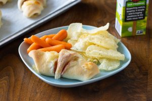 Ham and cheese rolled up in Rhodes dough. Roll up is on a plate with chips and carrots.
